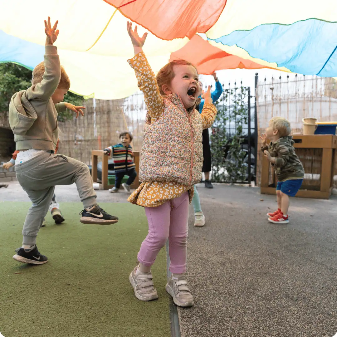 Children enjoying outdoor play under a colorful parachute at nursery.