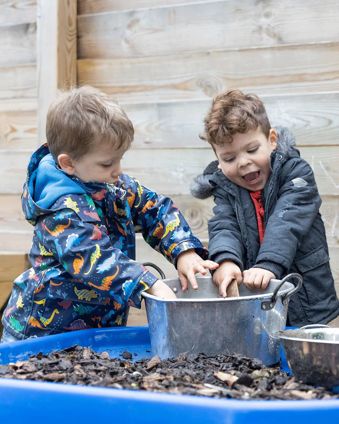 Two young children playing with soil and water in an outdoor nursery setting.